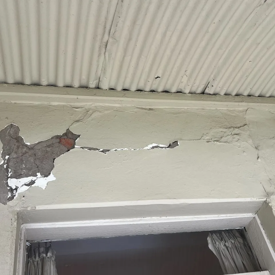 Verandah wall under a corrugated-iron ceiling, render blown off in a patch down to the brick, a crack running above the window head